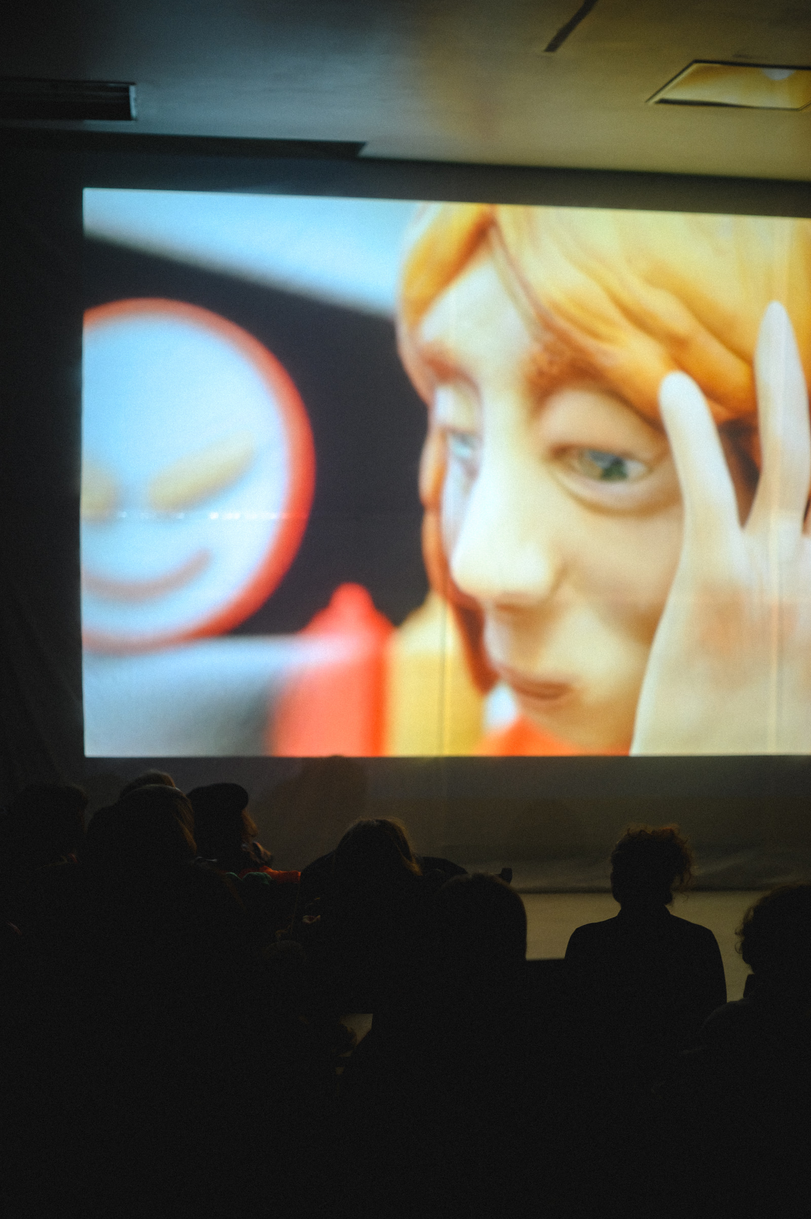 A large screen displays a close-up of an animated character with light-colored hair and expressive eyes, while a group of people sit in front of it watching.
