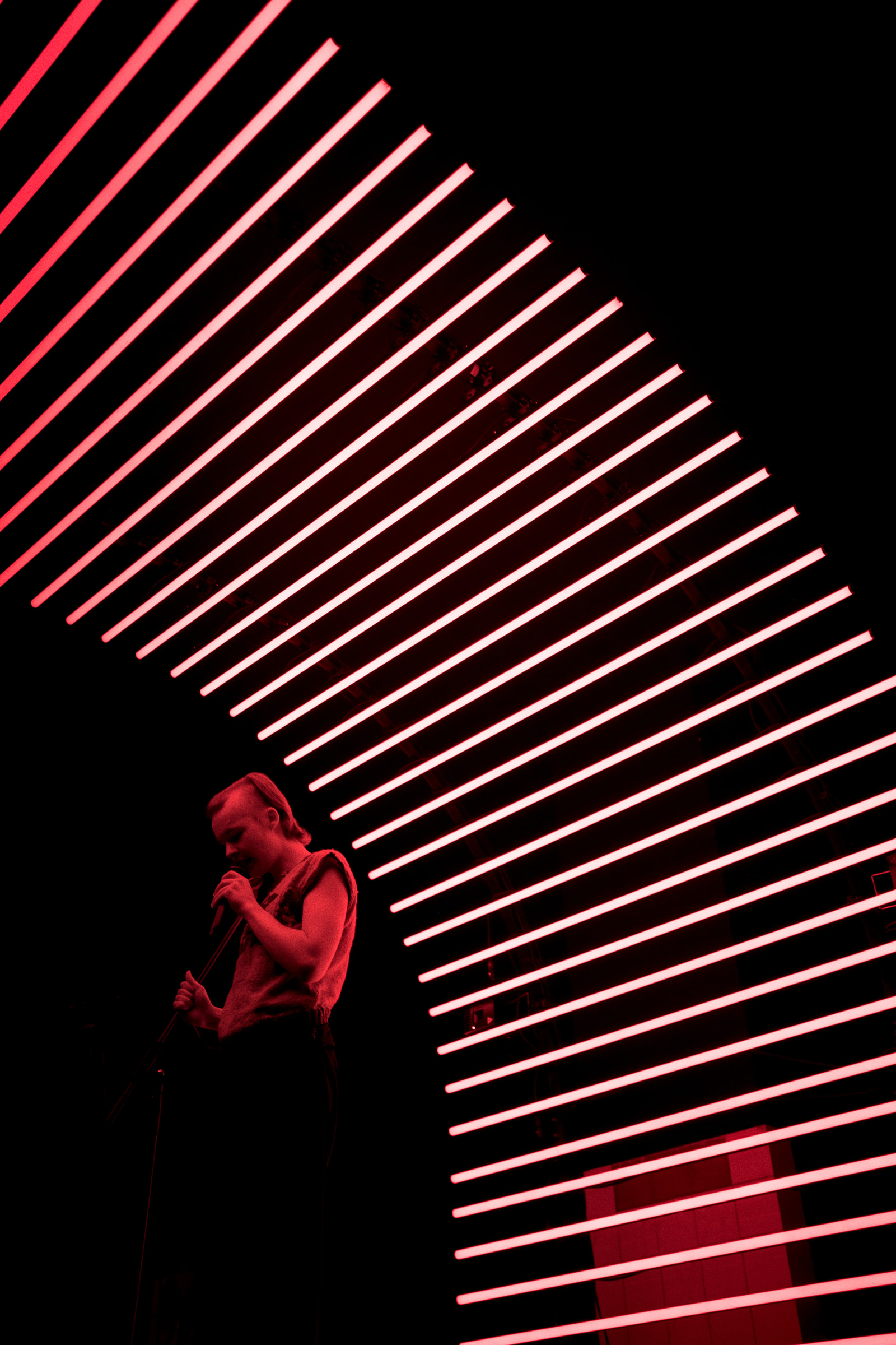 A person sings into a microphone against a backdrop of vertical red neon lights.