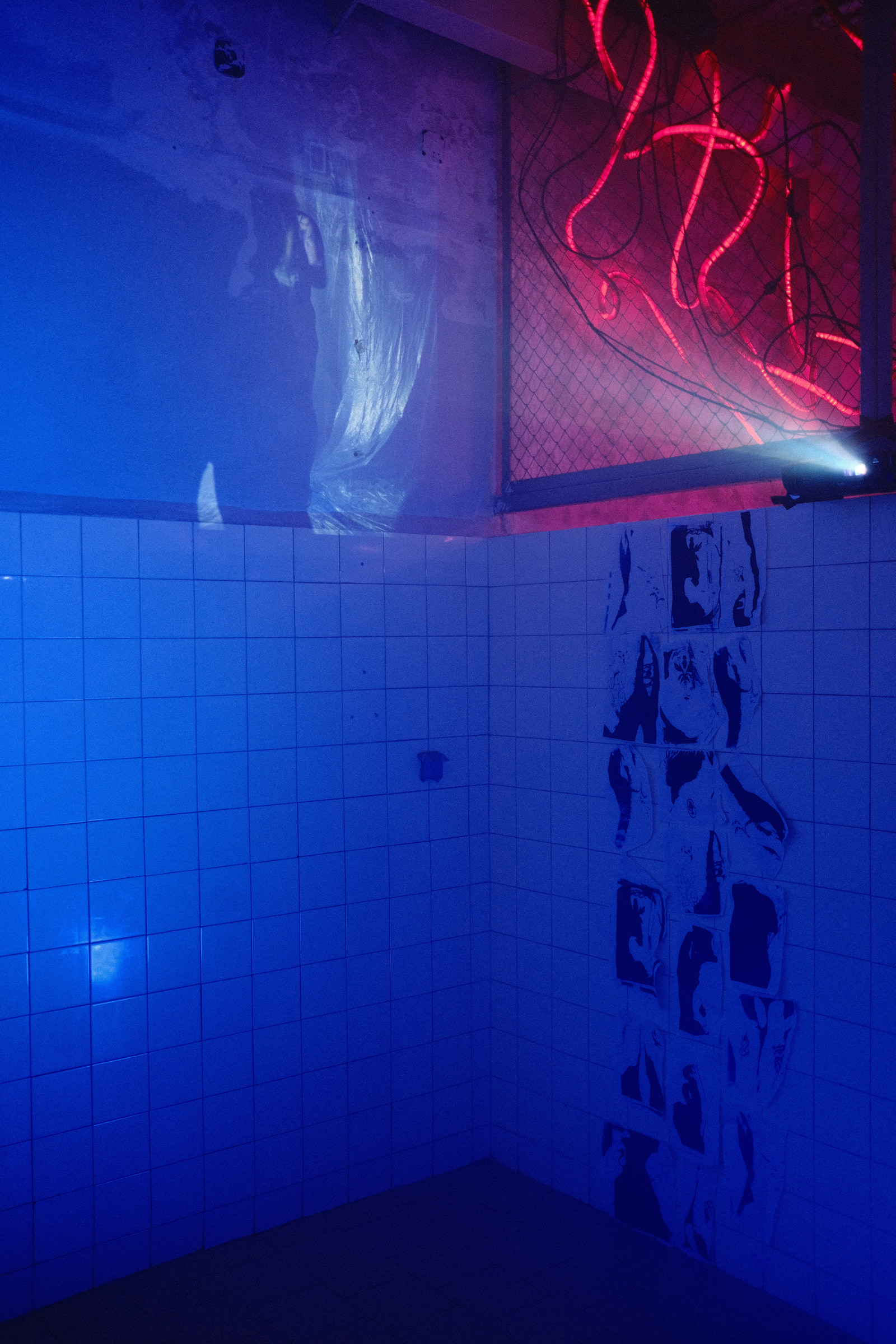 A small room with tiled walls and floor, illuminated by blue lighting, features a neon sign with red lettering and a draped fabric on one wall.