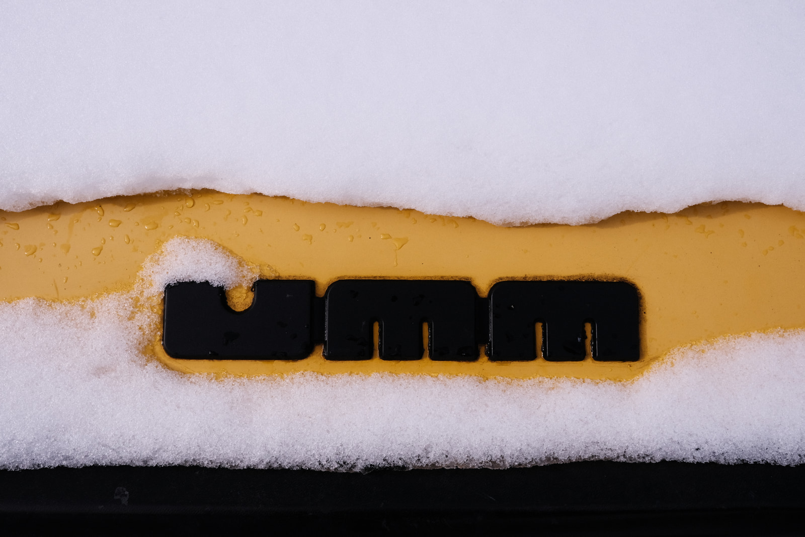 Black logo with three letters on a yellow background partially covered by snow.