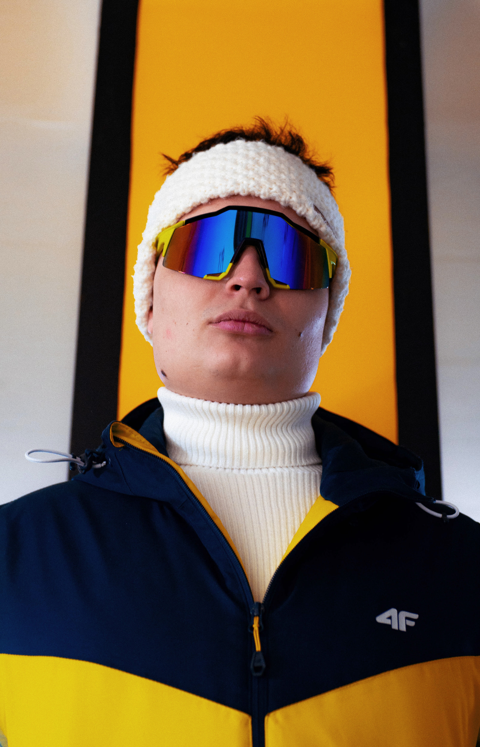 A person in a white beanie and reflective sunglasses wears a dark blue and yellow jacket against a bright orange background.
