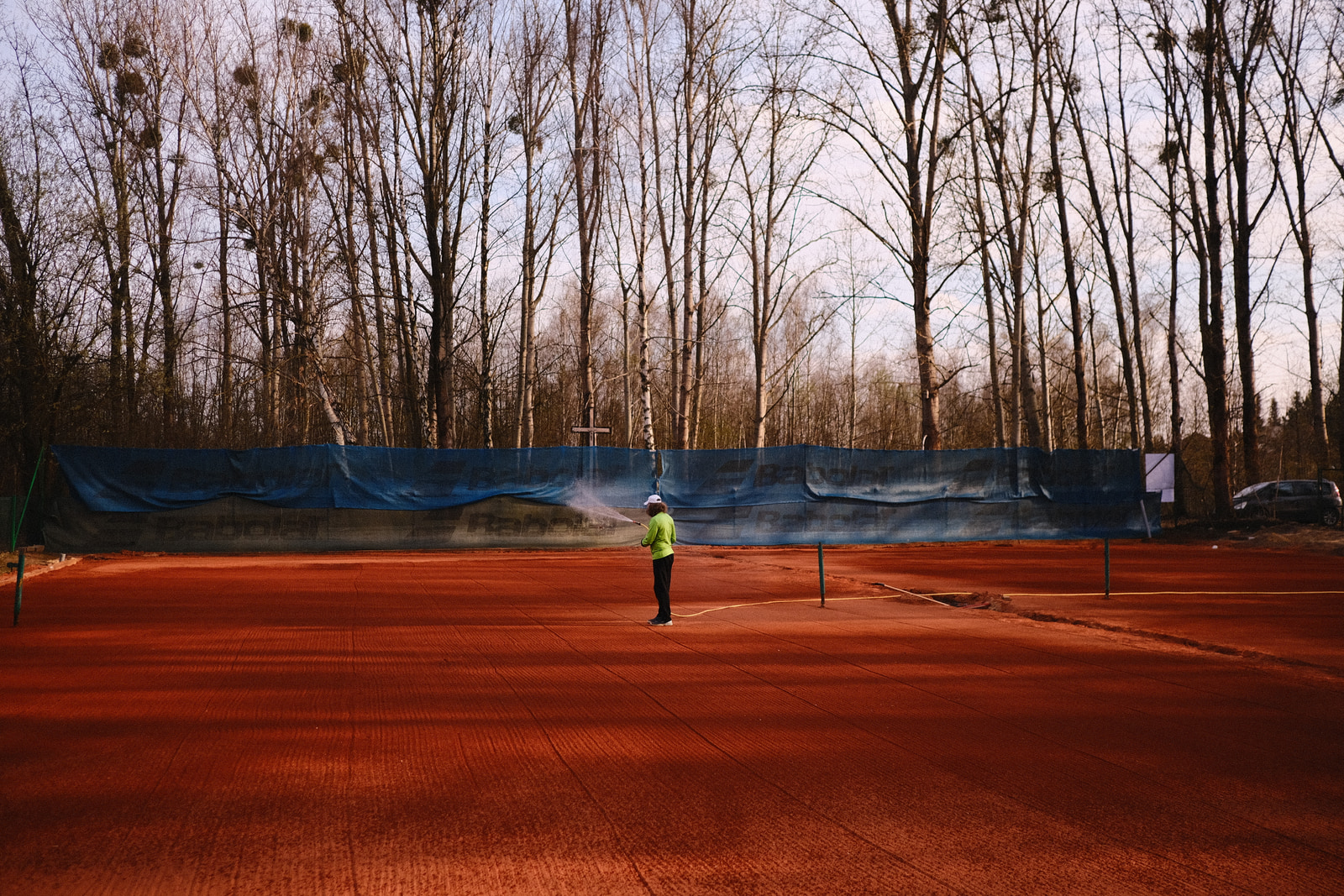 A person stands on a reddish-brown tennis court, with a blue net and leafless trees in the background.