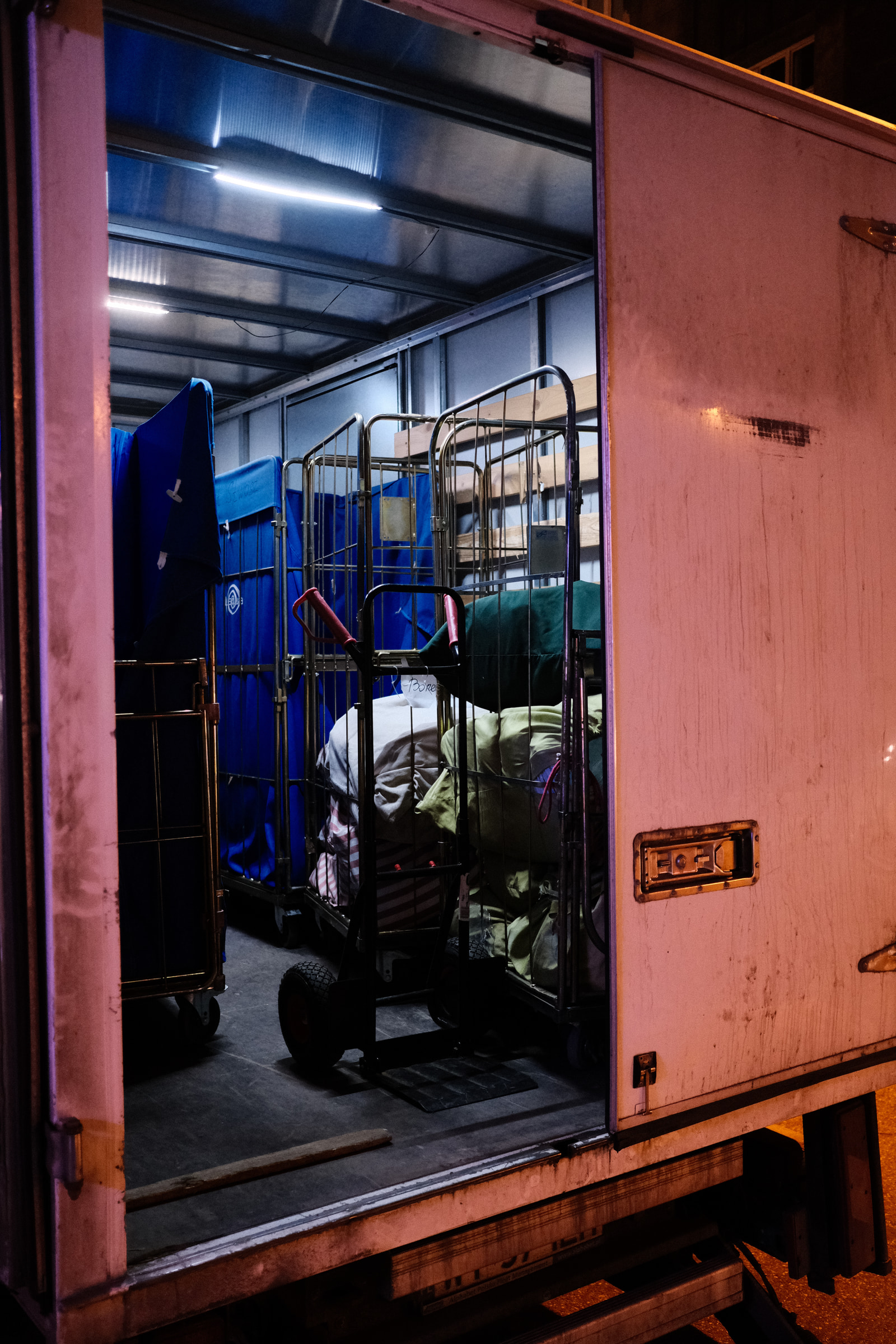An open trailer at night contains stacked blue containers and folded mattresses on metal racks.