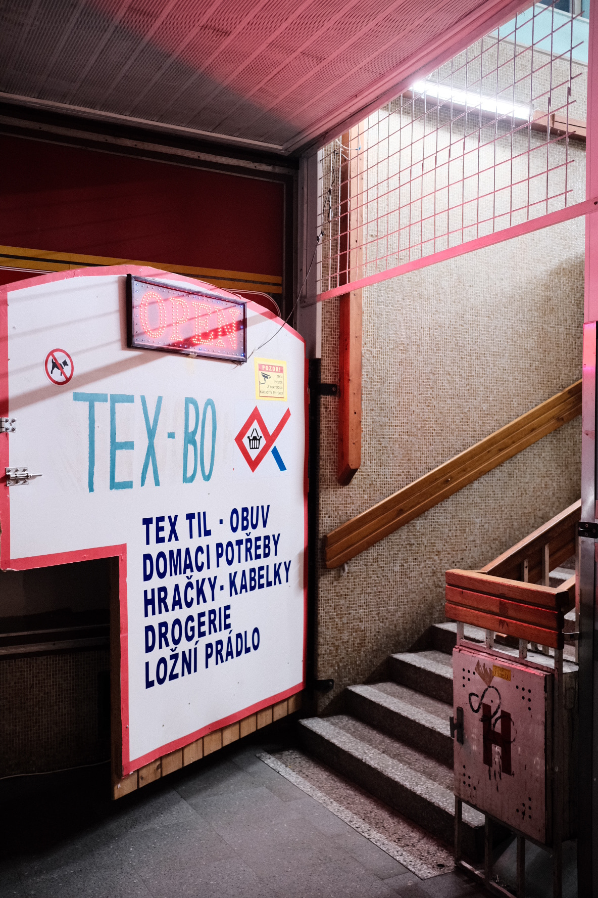 A signboard reading "TEX-BO" and additional text in Czech is mounted on a wall next to a staircase with wooden railings.