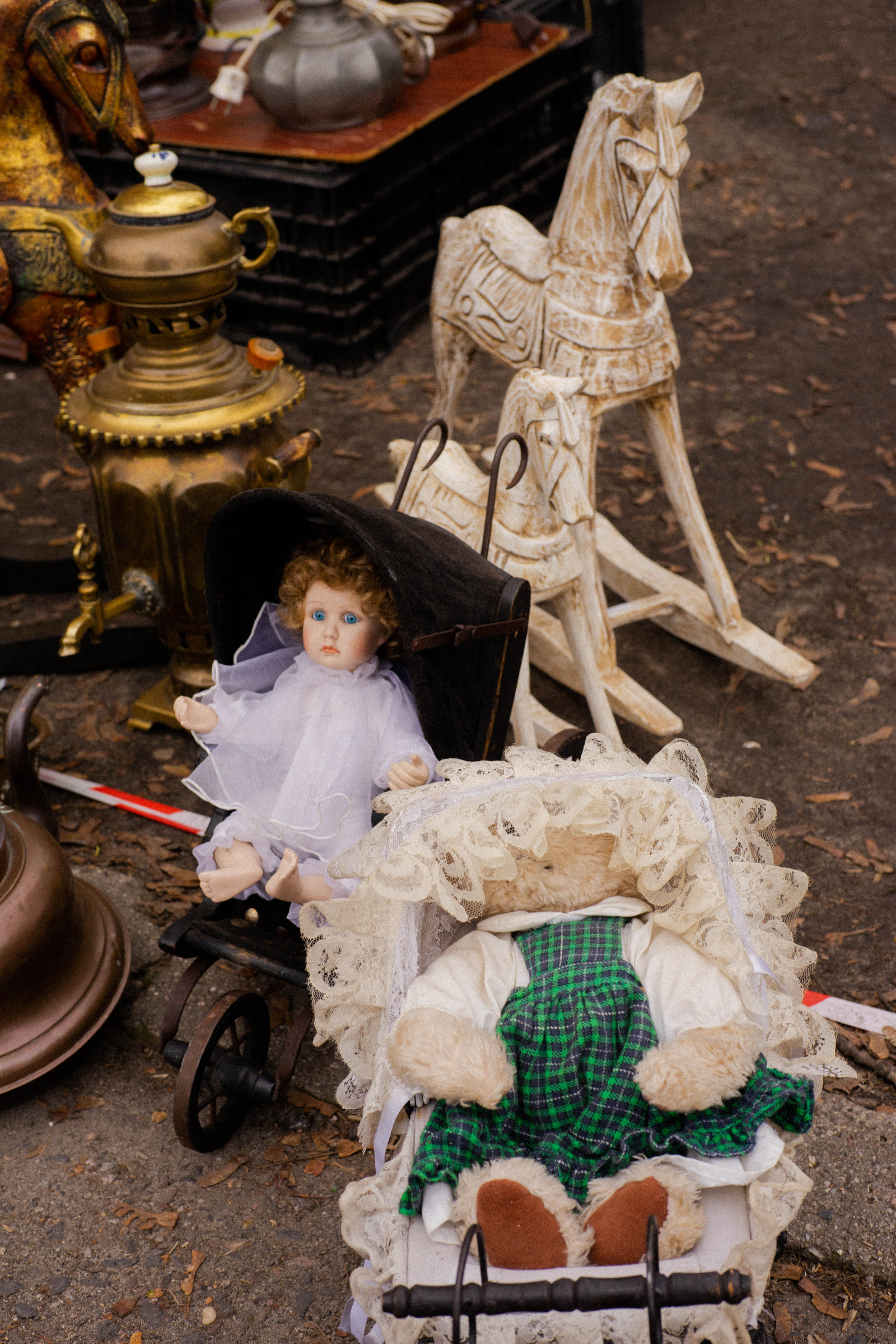 A vintage doll in a purple dress sits in a black pram next to a teddy bear in a green plaid outfit, surrounded by antique teapots and a wooden rocking horse on a paved surface.