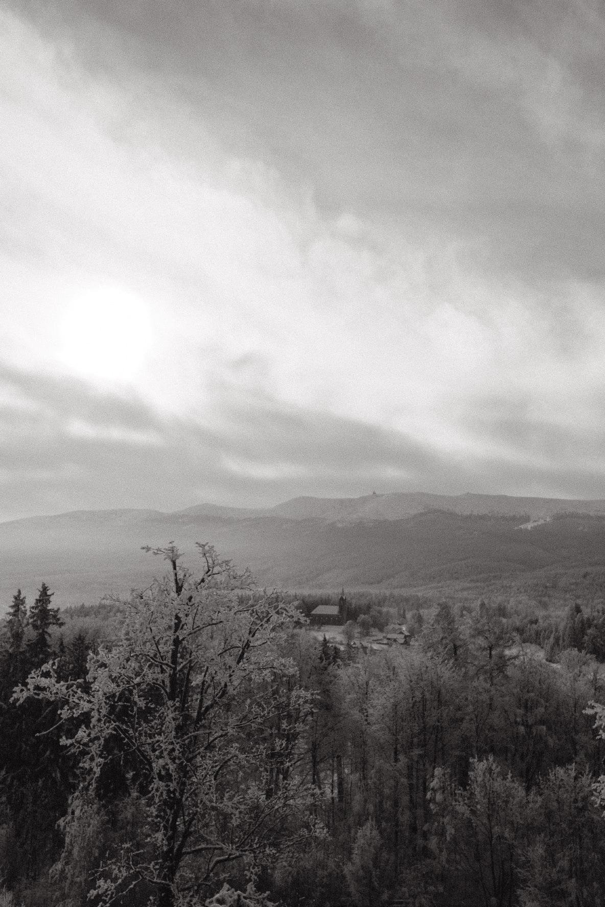 A wide, misty valley stretches beneath a cloudy sky, with a small settlement nestled among trees in the foreground.