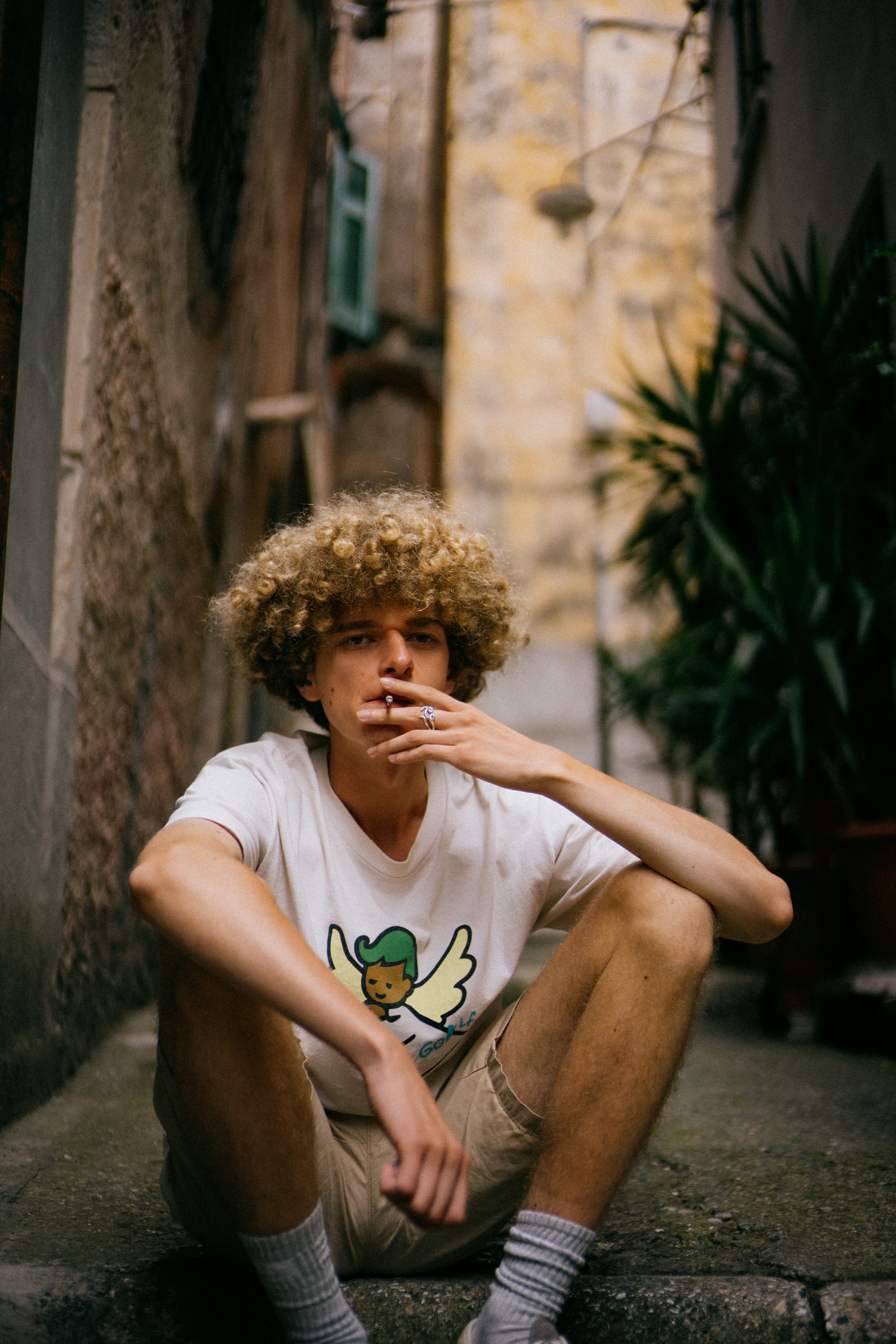 A young man sits on a stone step in a narrow alleyway, smoking a cigarette with one hand resting on his knee.