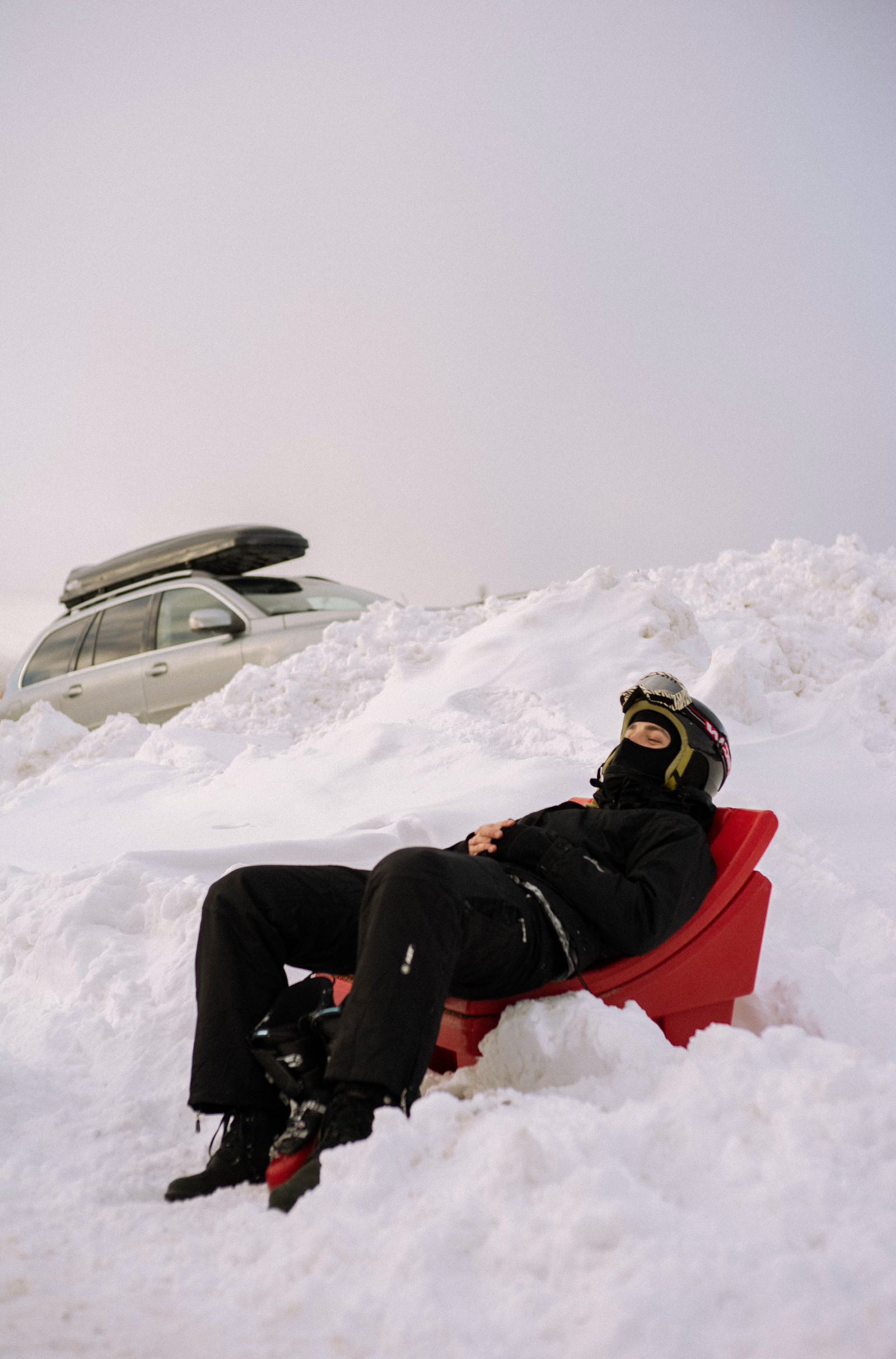 A person lies on a red lounge chair in deep snow, wearing a helmet and goggles, with a vehicle partially buried in the snow behind them.
